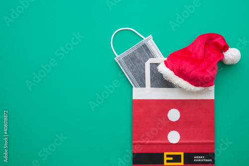 Covid-19 in Christmas festival celebration concept. Flat lay of red santa claus hat and protective face mask in gift bag on green background with copy space. photo