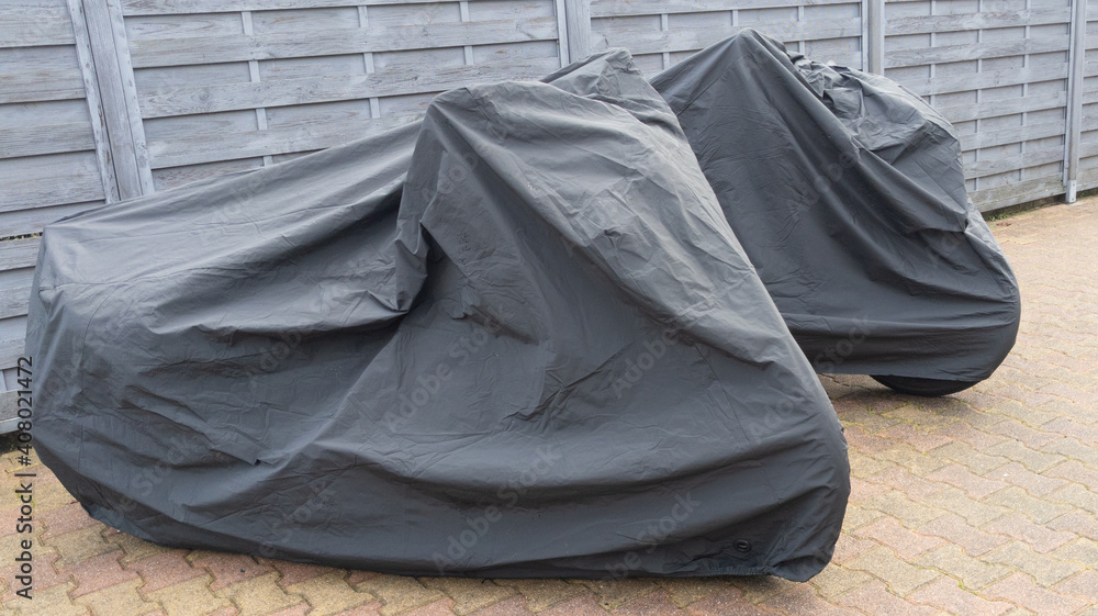 two motorcycles protected by black grey protective cover in city street motorbike with tarpaulin jacket two motorcycles protected by black grey protective cover in city street motorbike with tarpaulin jacket