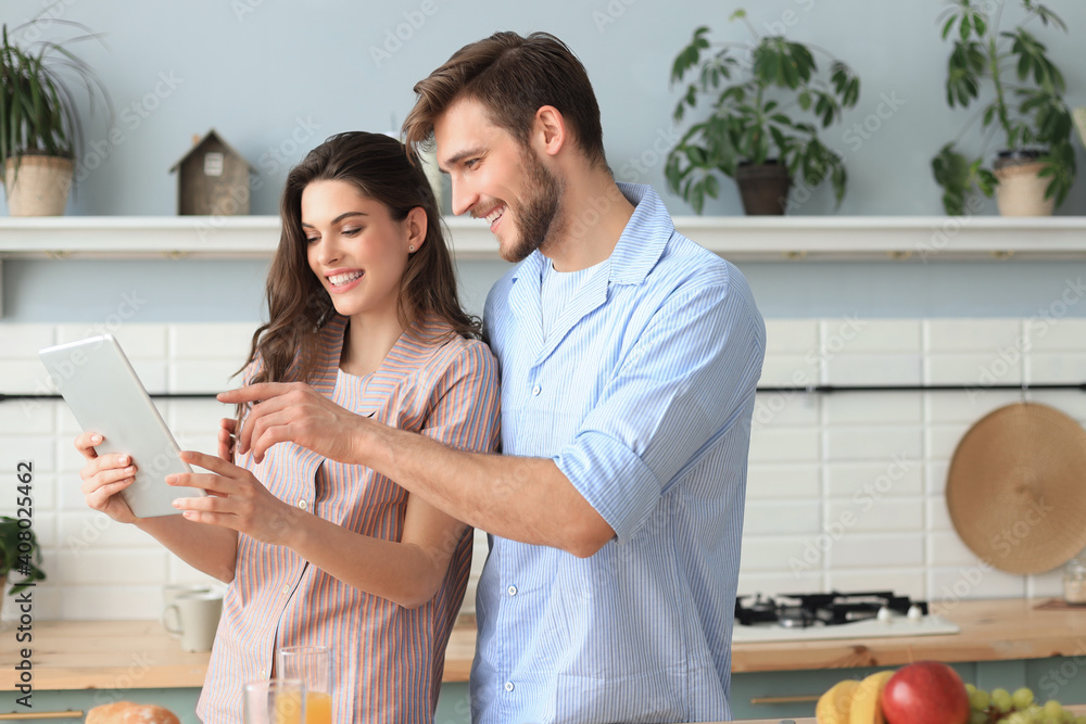 Happy young couple in pajamas watching online content in a didital tablet and smiling in kitchen at home. Happy young couple in pajamas watching online content in a didital tablet and smiling in kitchen at home.