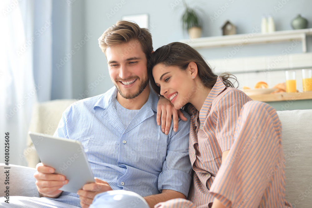 Young couple in pajamas watching media content online in a tablet sitting on a sofa in the living room. - obrazy, fototapety, plakaty 