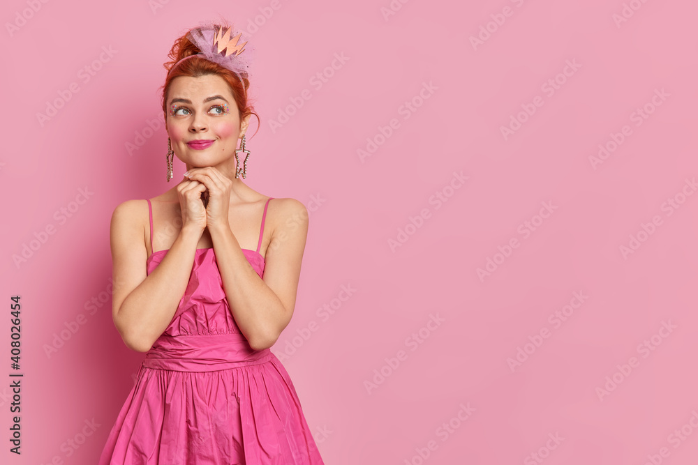 Studio shot of thoughtful dreamy expression has bright makeup keeps hands under chin prepares for carnival party wears crown and pink dress stands indoor with blank space for your promotion. Studio shot of thoughtful dreamy expression has bright makeup keeps hands under chin prepares for carnival party wears crown and pink dress stands indoor with blank space for your promotion.