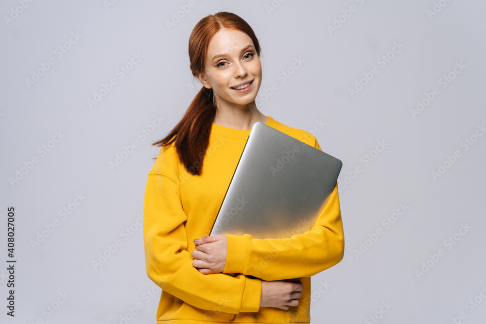 Obraz premium Cheerful young woman student holding closed laptop computer and looking at camera on isolated white background. Pretty lady model with red hair emotionally showing facial expressions in studio.