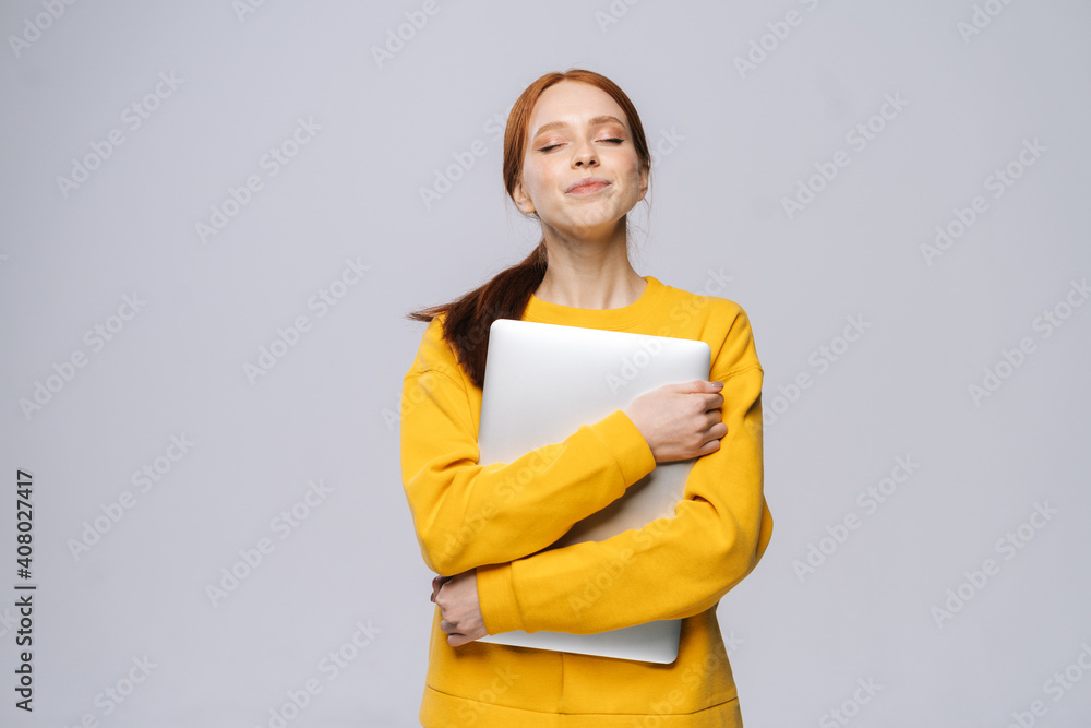 Obraz premium Happy charming young woman student holding laptop computer with closed eyes on isolated gray background. Pretty lady model with red hair emotionally showing facial expressions in studio, copy space.