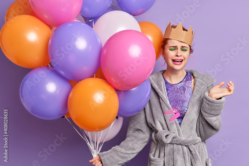 Upset redhaired woman with bright makeup cries from despair feels lonely on party holds balloons has spoiled birthday celebration wears paper crown and dressing gown isolated over purple background