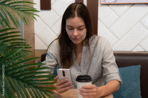 young woman sits at a table in a cafe and communicates on the Internet in mobile apps, smartphone and takeaway coffee in her hand