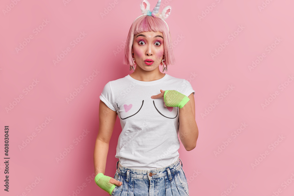 Surprised pink haired young Asian woman indicates at herself asks who me has puzzled expression dressed in fashionable clothes shocked to be mentioned by someone poses at studio. Do you mean me - obrazy, fototapety, plakaty 