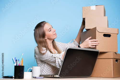 Small business, young female entrepreneur working at a Desk, using a laptop for work, managing an online store and communicating with customers. a stack of cardboard boxes is ready for delivery