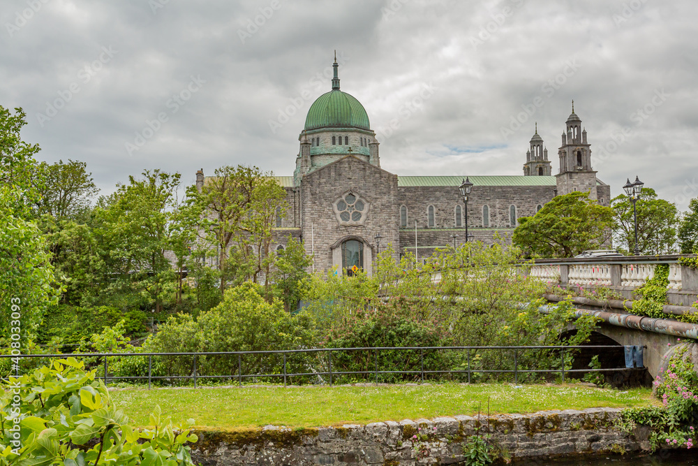 Cathedral of Our Lady Assumed into Heaven and St Nicholas with its green dome surrounded by green vegetation, Salmon Weir bridge over the Corrib river, cloudy day in Galway, Connacht province, Ireland