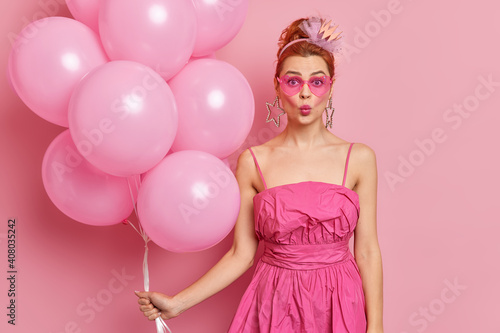 Photo of glamour woman likes pink color wears trendy sunglasses fashionable dress prefers style of nineties keeps lips rounded poses with inflated balloons celebrates holiday. Party concept.