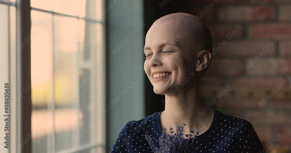 Head shot happy bald female cancer patient with lavender flowers bunch pose indoor smiles looks at camera feel alive, victory over oncology disease, remission, full recovery. World Cancer Day concept Head shot happy bald female cancer patient with lavender flowers bunch pose indoor smiles looks at camera feel alive, victory over oncology disease, remission, full recovery. World Cancer Day concept