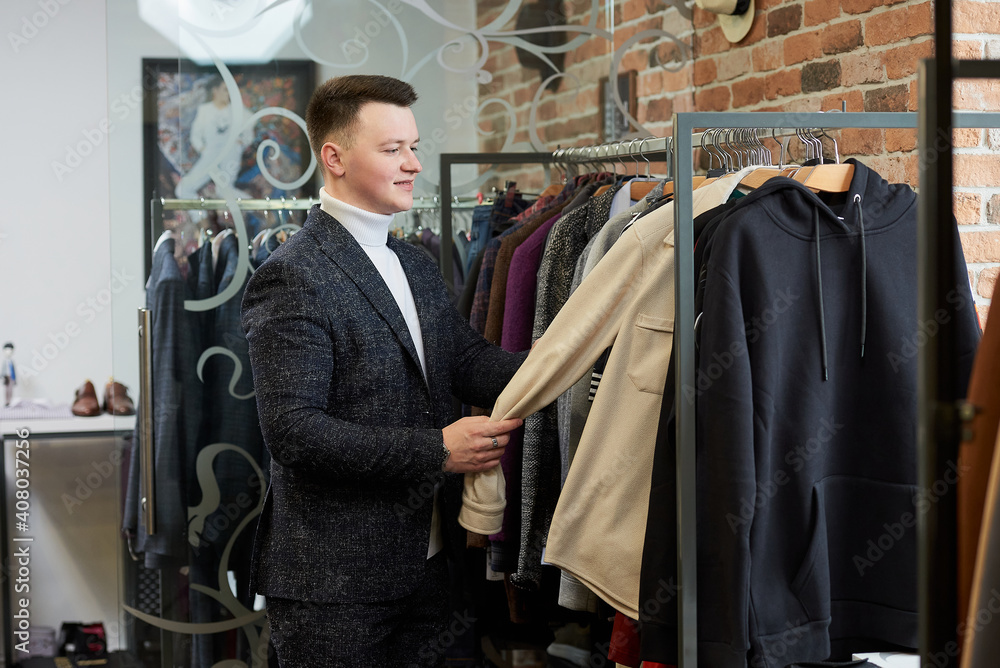 A young stylish man in a suit is choosing a shirt or hoodie in a clothing store. A male customer in a boutique. A young stylish man in a suit is choosing a shirt or hoodie in a clothing store. A male customer in a boutique.