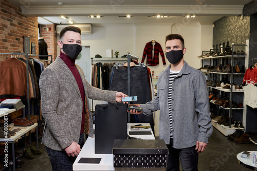 A man in a face mask is posing while using a smartphone to pay by NFC for purchases in a store. A shop assistant in a mask is holding out a terminal for contactless paying to a customer in a boutique