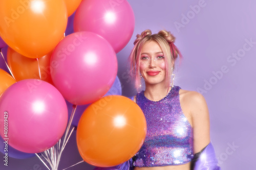 Slow glow effect. Pleased stylish blonde girl in eighties style poses at retro party with inflated colorful balloons poses against purple background. People partying celebration holiday concept