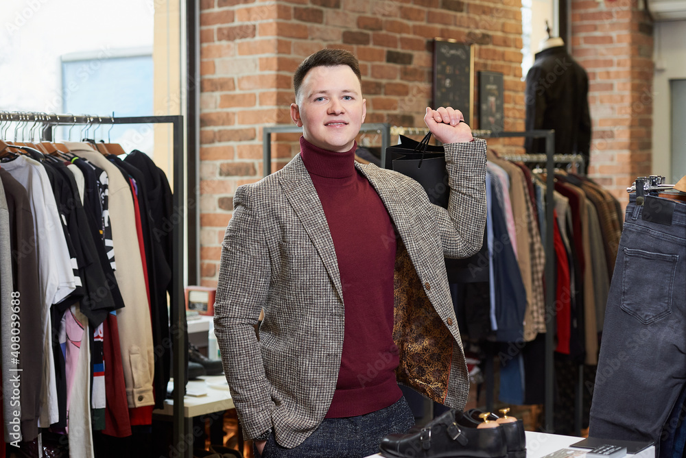 A young man in a sports jacket is posing with his purchases in black paper bags in a clothing store. A happy male customer in a boutique. A young man in a sports jacket is posing with his purchases in black paper bags in a clothing store. A happy male customer in a boutique.