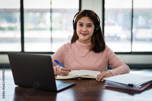 Smiling Asian girl student wears wireless headphones write on the notebook to study language online watch and listen to the lecturer  webinar via video call e-learning at home  distance education