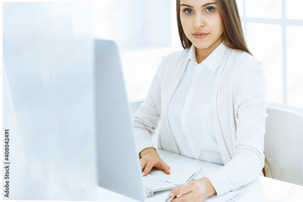 Young cheerful woman sitting at the desk with computer and looking at camera in white colored office. Looks like student girl or business lady. Headshot Young cheerful woman sitting at the desk with computer and looking at camera in white colored office. Looks like student girl or business lady. Headshot