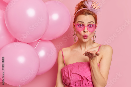 Romantic redhead woman sends mwah at camera expresses love to beloved person wears trendy pink sunglasses and dress poses with inflated balloons at birthday party. People special holiday concept