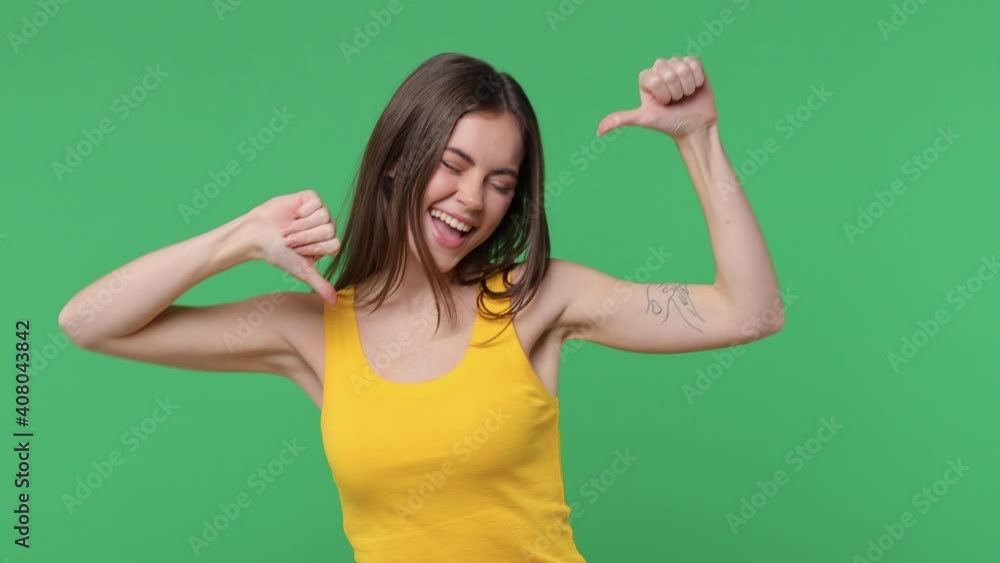 Funny brunette young woman 20s in yellow tank top posing isolated on green background in studio. People emotions lifestyle concept. Pointing fingers on herself blinking showing thumb up like gesture Funny brunette young woman 20s in yellow tank top posing isolated on green background in studio. People emotions lifestyle concept. Pointing fingers on herself blinking showing thumb up like gesture