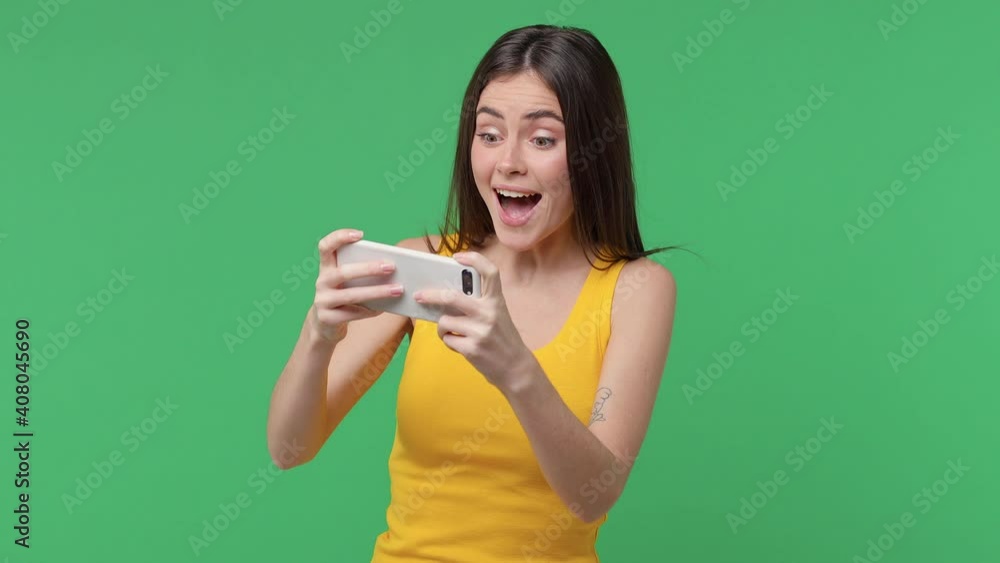 Excited happy brunette young woman 20s years old in yellow tank top posing isolated on green background studio. People lifestyle concept. Play game on mobile cell phone doing winner gesture say yes Excited happy brunette young woman 20s years old in yellow tank top posing isolated on green background studio. People lifestyle concept. Play game on mobile cell phone doing winner gesture say yes
