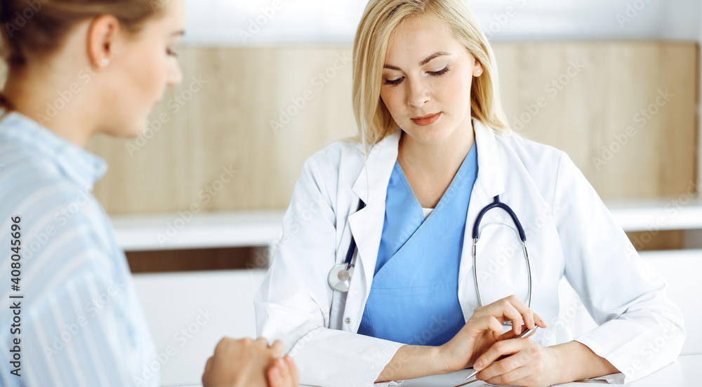 Woman-doctor and patient sitting and talking in hospital or clinic. Blonde therapist is cheerfully smiling. Medicine concept Woman-doctor and patient sitting and talking in hospital or clinic. Blonde therapist is cheerfully smiling. Medicine concept