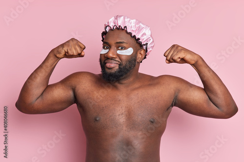 Self confident bearded black man raises arms brags his muscles wears beauty patches and bath hat looks seriously at camera going to take shower isolated over pink background. Hygiene and skin care