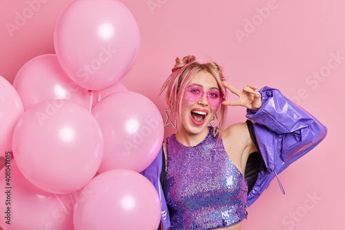 Photo of carefree joyful woman has fun on birthday party wears trendy sunglasses and purple jacket exclaims with happiness makes peace gesture holds bunch of inflated balloons poses over pink wall