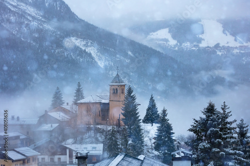 Church in Champagny-en-Vanoise village in France and view on Courchevel during heavy snowfall at winter