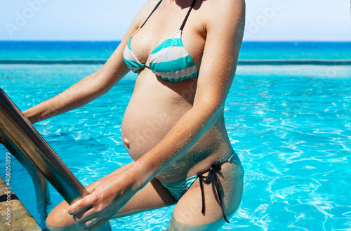 Close-up photo of a pregnant woman enter swimming pool with sea view holding rails belly close up