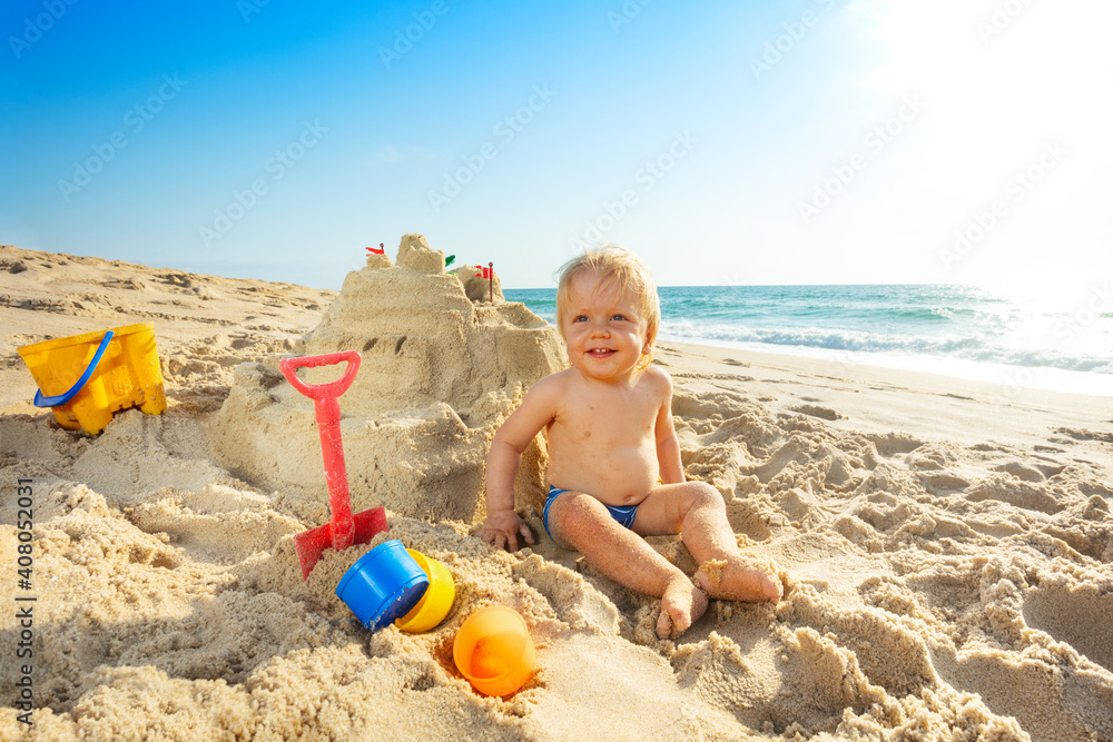 Happy cute little toddler boy sit near sand castle with plastic bucket shovel and other toys smiling Happy cute little toddler boy sit near sand castle with plastic bucket shovel and other toys smiling