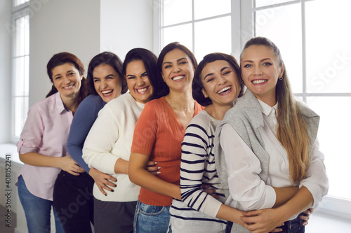Group portrait of female friends hugging, smiling and looking at camera. Team of happy young women in their 20s and 30s standing in bright office and embracing each other. Concept of unity and support
