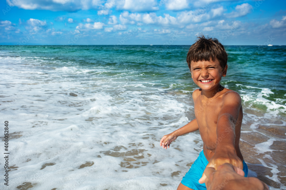 Happy little boy smiling pull parent's hand to the sea with big smile and waves on background Happy little boy smiling pull parent's hand to the sea with big smile and waves on background