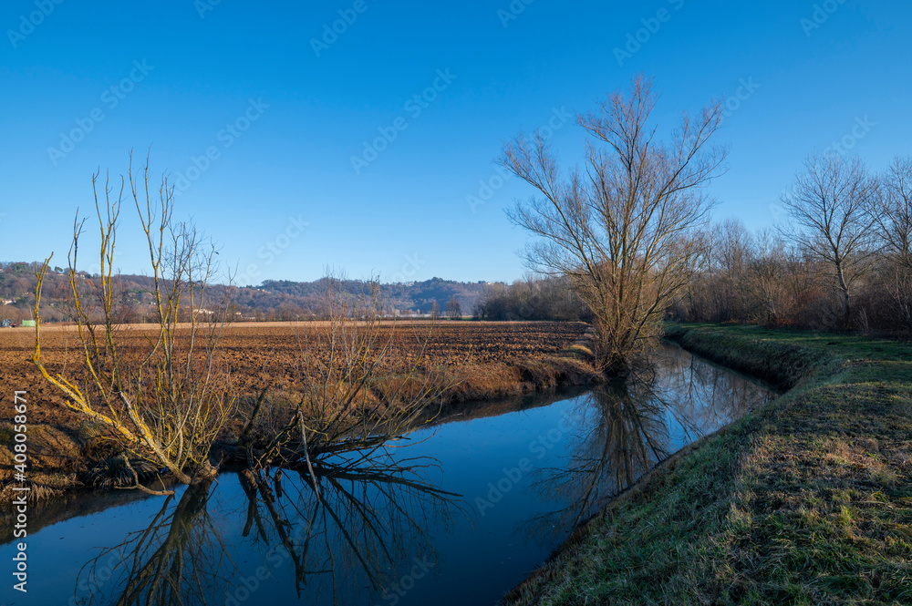 Panorama of the northern Italian countryside, detail of the river and the embankments where the trees are reflected in the river with the reflections of the sun on an autumn day, cold but with the sun Panorama of the northern Italian countryside, detail of the river and the embankments where the trees are reflected in the river with the reflections of the sun on an autumn day, cold but with the sun