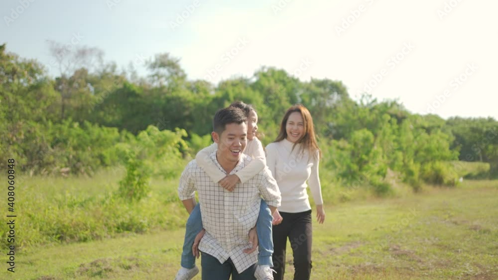 Happy young asian father piggyback with his little girl while mother run to play with her in the park and enjoyed ourselves together in the green garden. Family enjoying sunny fall day in nature. Happy young asian father piggyback with his little girl while mother run to play with her in the park and enjoyed ourselves together in the green garden. Family enjoying sunny fall day in nature.