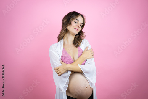 Young beautiful brunette woman pregnant expecting baby over isolated pink background hugging oneself happy and positive, smiling confident