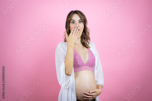 Young beautiful brunette woman pregnant expecting baby over isolated pink background covering mouth with hand, shocked and afraid for mistake. surprised expression