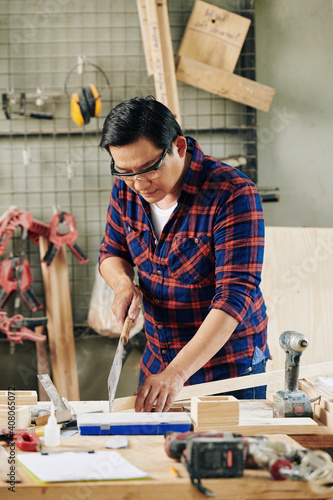 Mature experienced carpenter in plastic goggles sawing wooden planks