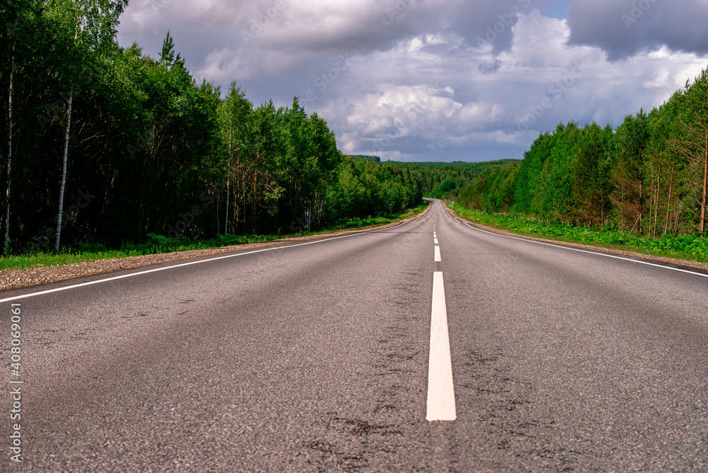 An asphalted intercity road with white markings extending into the distance through a green forest. An asphalted intercity road with white markings extending into the distance through a green forest.