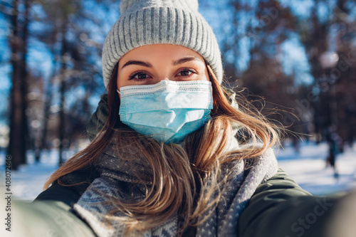 Woman wearing protective face mask in snowy winter park taking selfie. Corona virus covid-19 pandemic safety measures.