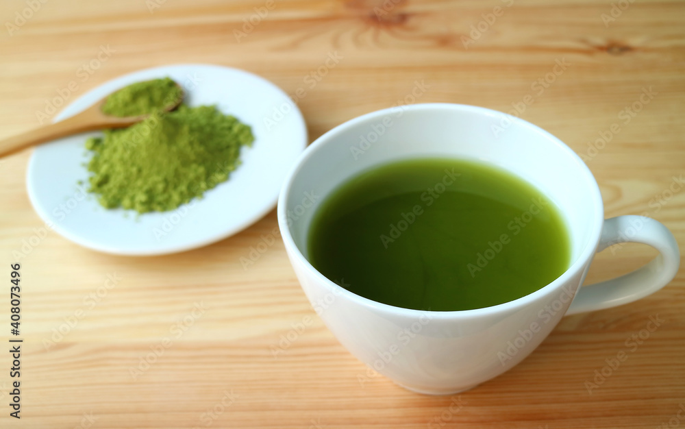 Closeup a Cup of Hot Matcha Green Tea on Wooden Table with Blurry Matcha Tea Powder in Background Closeup a Cup of Hot Matcha Green Tea on Wooden Table with Blurry Matcha Tea Powder in Background