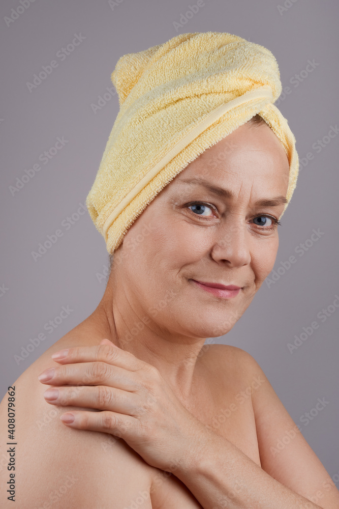 Smiling middle aged woman with bare shoulders applying cream to her skin, studio shot in profile on gray background. Facial skin care concept. Smiling middle aged woman with bare shoulders applying cream to her skin, studio shot in profile on gray background. Facial skin care concept.