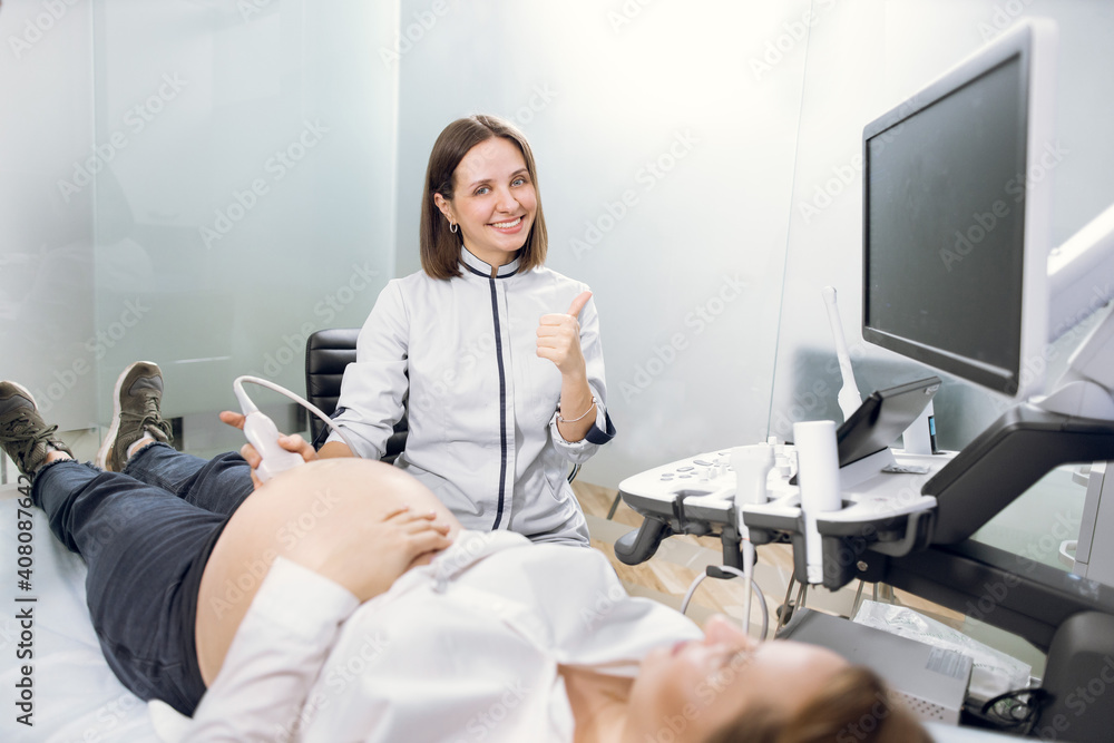 Obraz premium Portrait of young attractive female doctor, posing to camera with smile and thumb up, while conducting ultrasound scanning procedure for her patient, young pregnant woman. Ultrasound scan concept