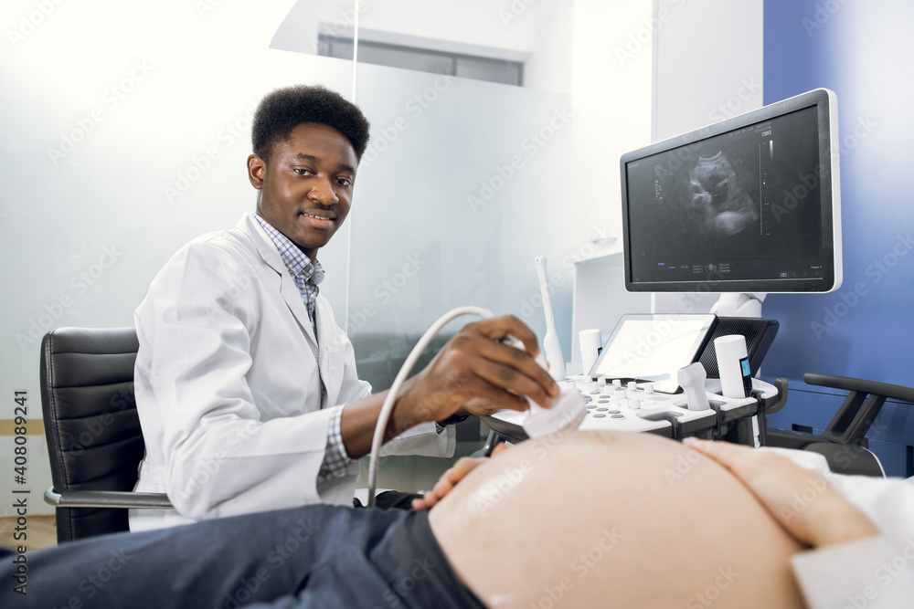 Smiling African American man doctor making the ultra sound of pregnant belly for female patient. Medical exam of a young pregnant woman by ultrasound equipment