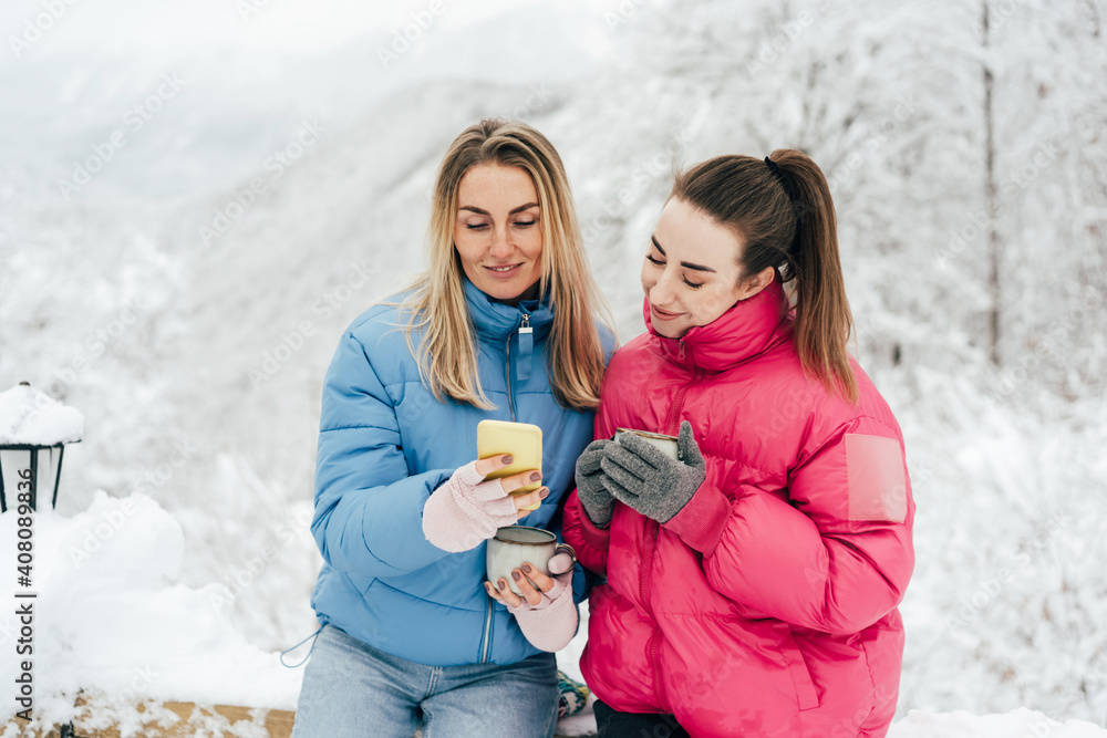 Obraz premium Two young women in bright jackets outdoors in snowy weather are watching social networks and photographs on the phone.