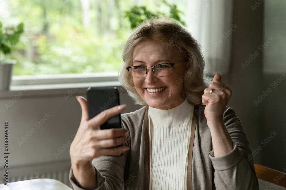 Close up overjoyed mature woman wearing glasses using phone, celebrating success, showing yes gesture, reading good news in message, received great shopping offer, looking at smartphone screen