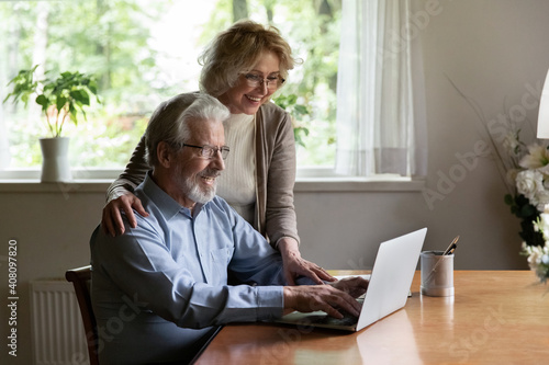 Happy mature man and woman wearing glasses using laptop together, chatting with relatives or shopping online, smiling elderly wife and husband looking at computer screen, having fun with device