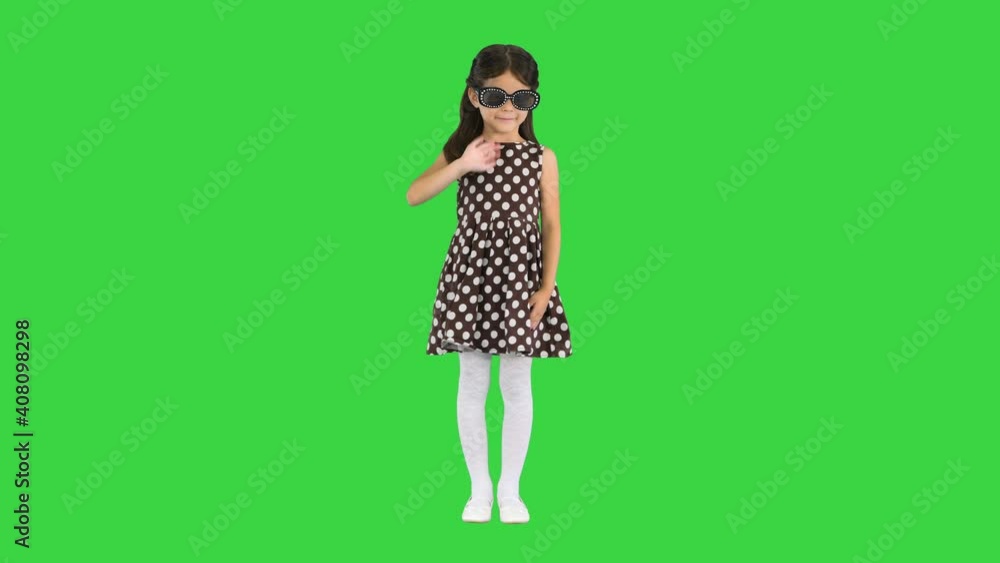 Dark-haired little girl in polka-dot dress putting on black sun glasses and waving to the camera on a Green Screen, Chroma Key. Dark-haired little girl in polka-dot dress putting on black sun glasses and waving to the camera on a Green Screen, Chroma Key.