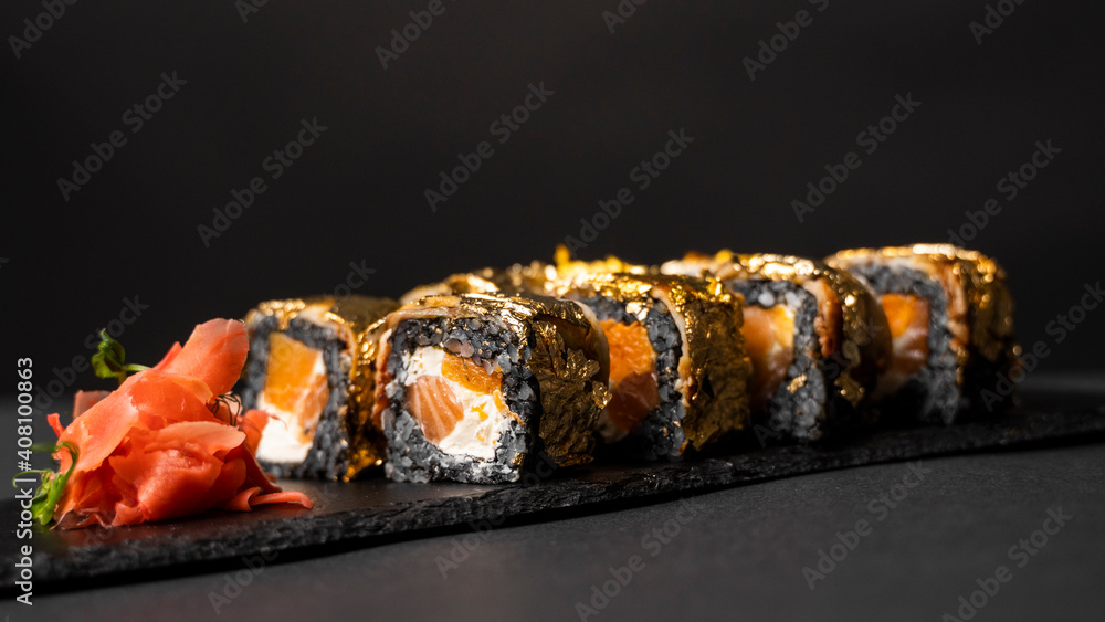 Custom sushi roll with nori, cuttlefish ink, fresh salmon, cream cheese, pepper pumpkin, eel, unagi sauce and edible gold leaf with red ginger, wasabi on a square plate on black table and background. Custom sushi roll with nori, cuttlefish ink, fresh salmon, cream cheese, pepper pumpkin, eel, unagi sauce and edible gold leaf with red ginger, wasabi on a square plate on black table and background.