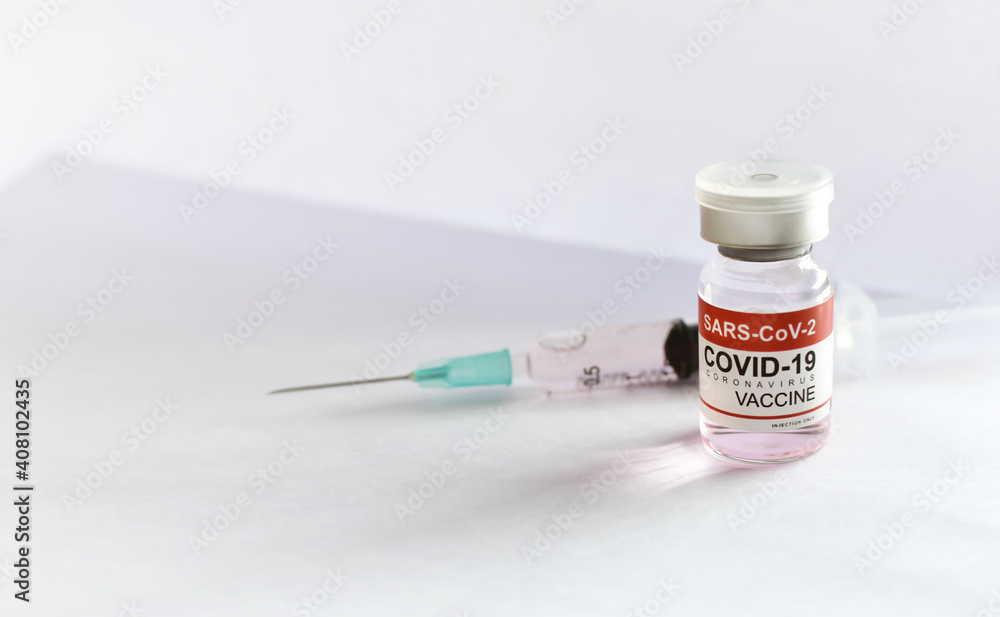 COVID-19 (SARS-CoV-2) coronavirus vaccine bottle with blurred syringe and white paper background COVID-19 (SARS-CoV-2) coronavirus vaccine bottle with blurred syringe and white paper background