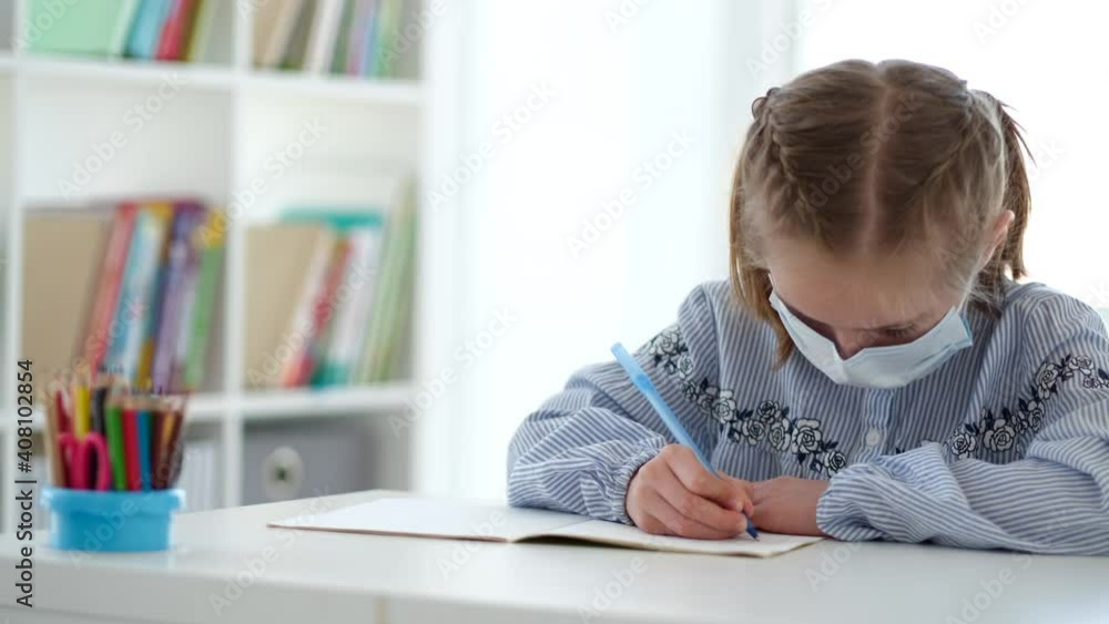 Little girl in protective mask writing in copybook with pen during lesson at school Little girl in protective mask writing in copybook with pen during lesson at school