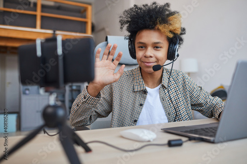 Portrait of teenage African-American boy wearing headset and waving at camera while streaming video games at home, young gamer or blogger concept, copy space photo